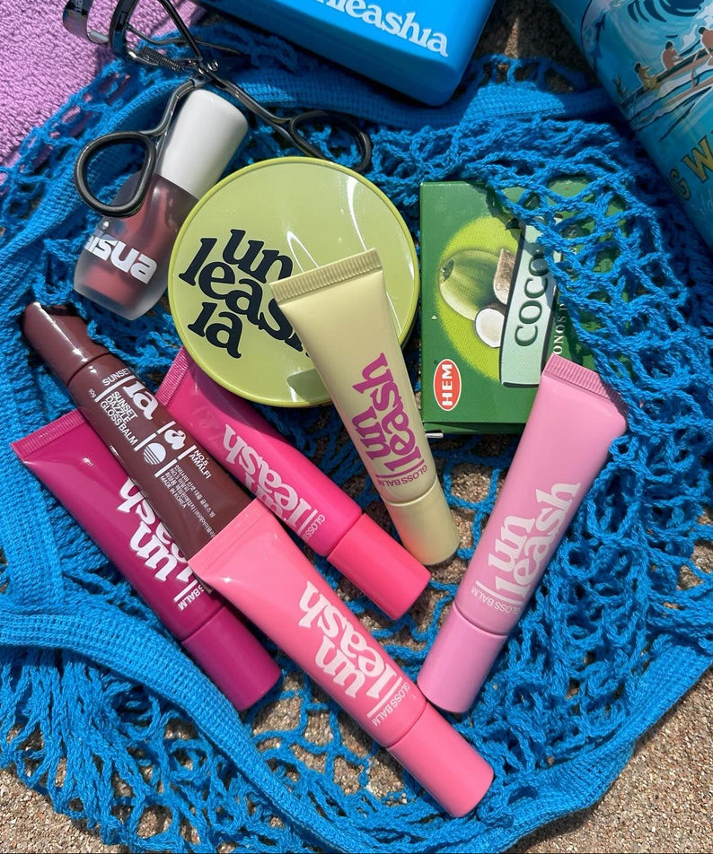 Various UNLEASHIA products including the UNLEASHIA Sunset Dazzle Gloss Balm displayed on a blue net.