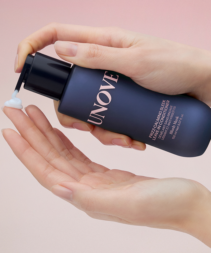 Hands holding a bottle of UNOVE Frizz Calming Sleek Leave-In Conditioner 150ml dispensing product, with a gradient background.