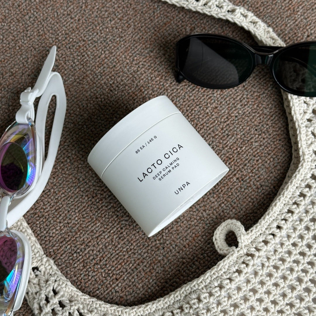 UNPA Lacto Cica Deep Calming Serum Pad 85 Sheets on a textured surface with sunglasses and a crochet bag.