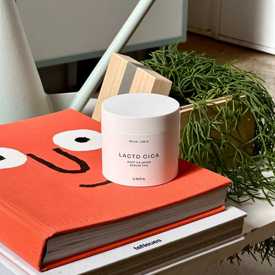 UNPA Lacto Cica Deep Calming Serum Pad 85 Sheets on an orange book beside green plant decor