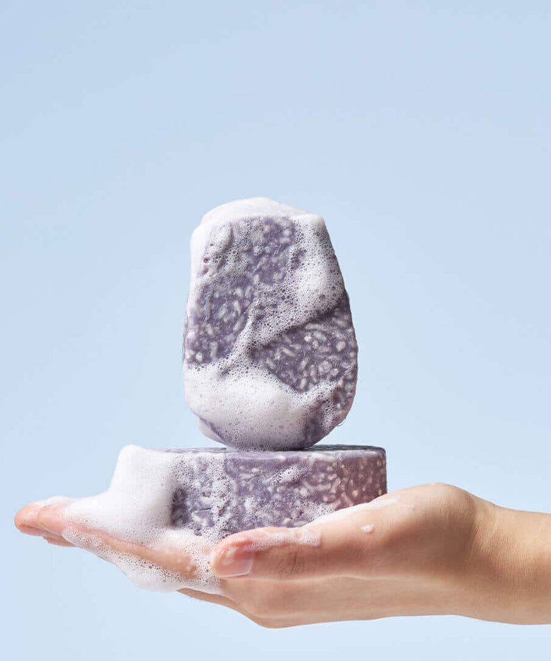 Hand holding two lathered URANG Daily Oil Free Shampoo Bars against a blue background.