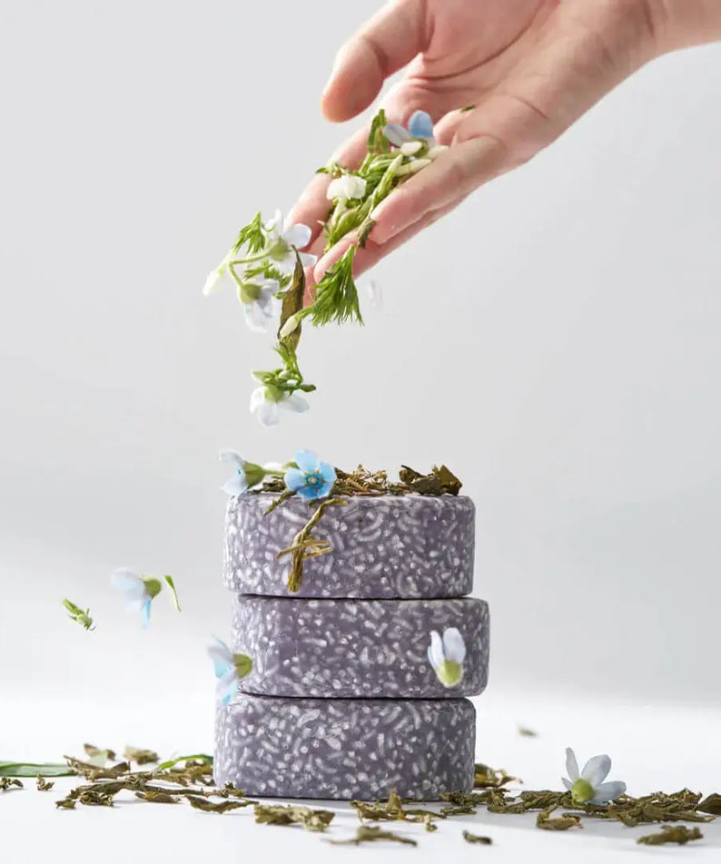 A hand decorating stack of URANG Daily Oil Free Shampoo Bar with flowers and leaves in a minimalist setting.