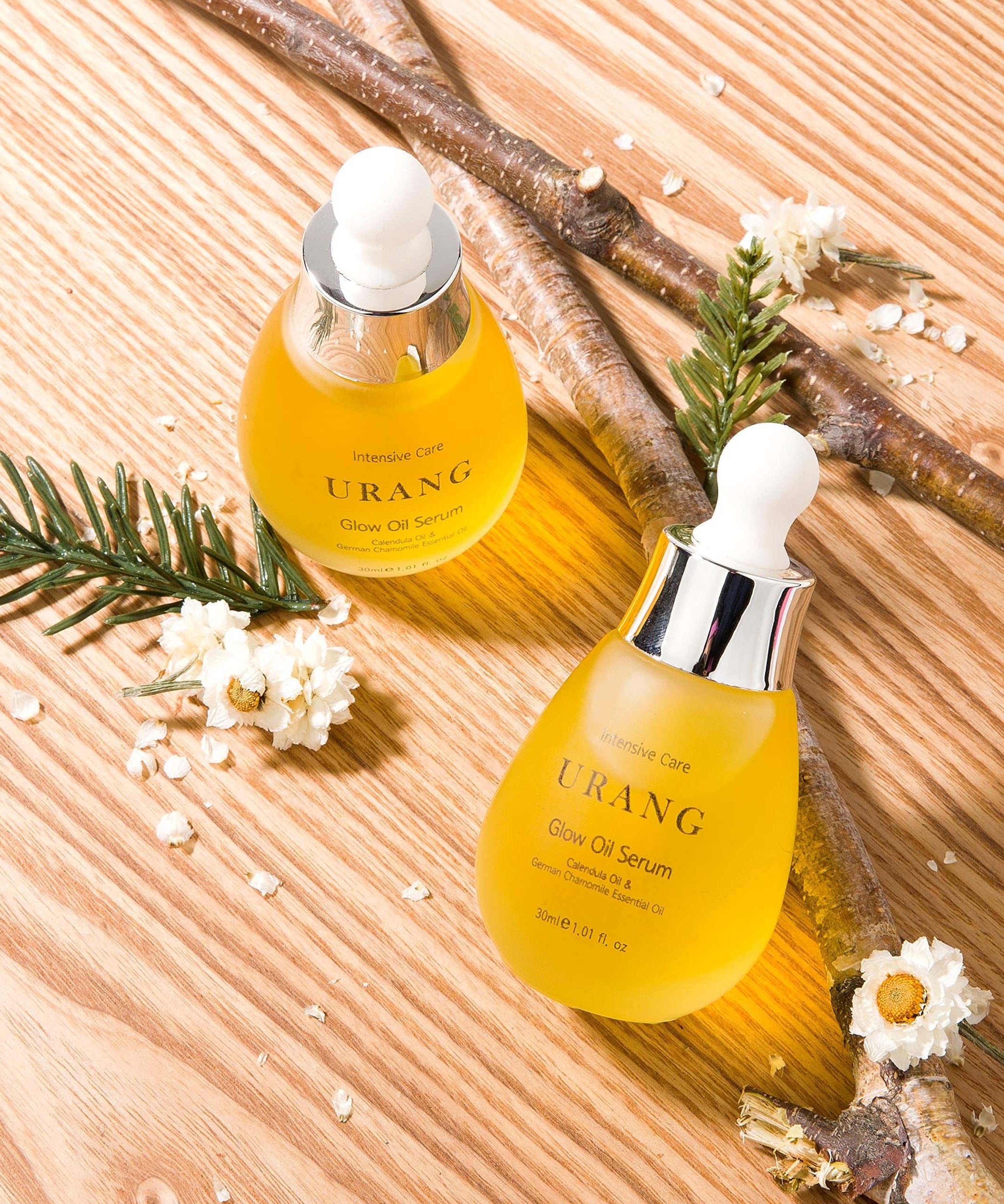 Two bottles of URANG Glow Oil Serum 30ml on wooden surface with flowers and twigs, showcasing skincare product.