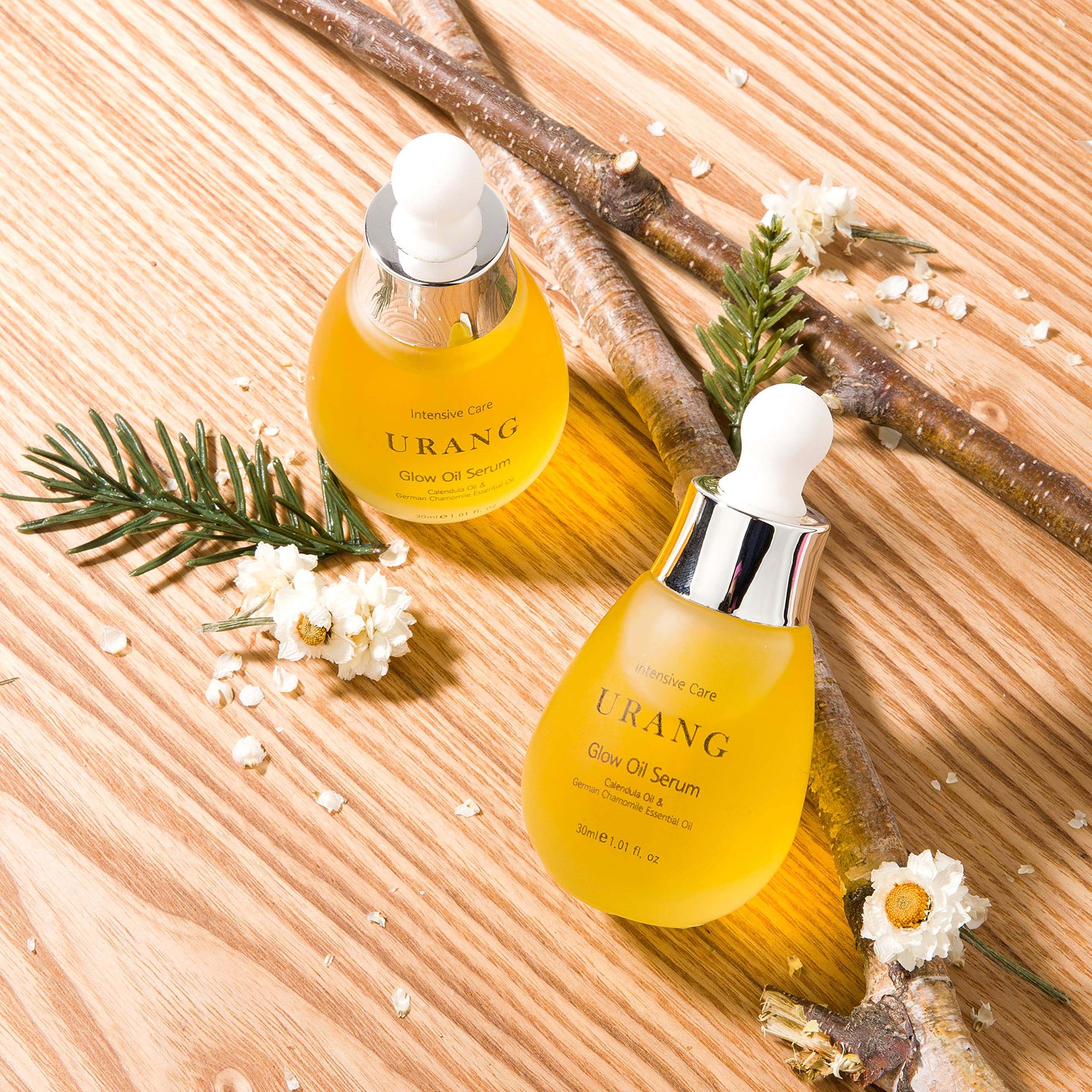 Two bottles of URANG Glow Oil Serum 30ml on wooden surface with flowers and twigs, showcasing skincare product.