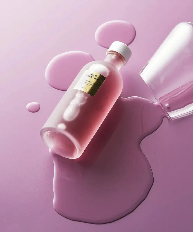 URANG Prestige Purple Shield Essence Toner bottle on spilled liquid, showcasing its luxurious rose-colored elixir on a purple surface.