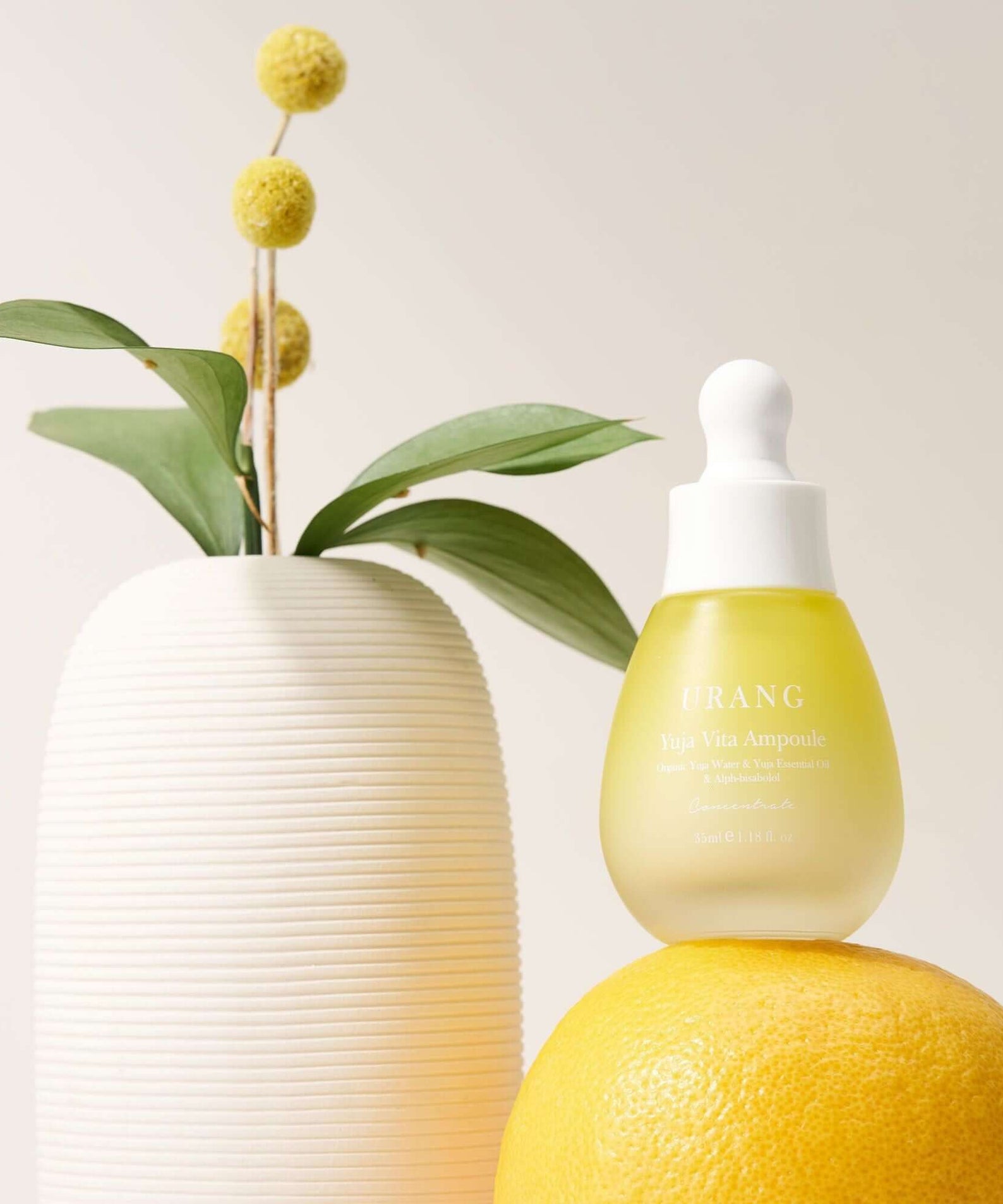 URANG Yuja Vita Ampoule bottle placed on a citrus fruit next to a modern vase with yellow flowers and green leaves.