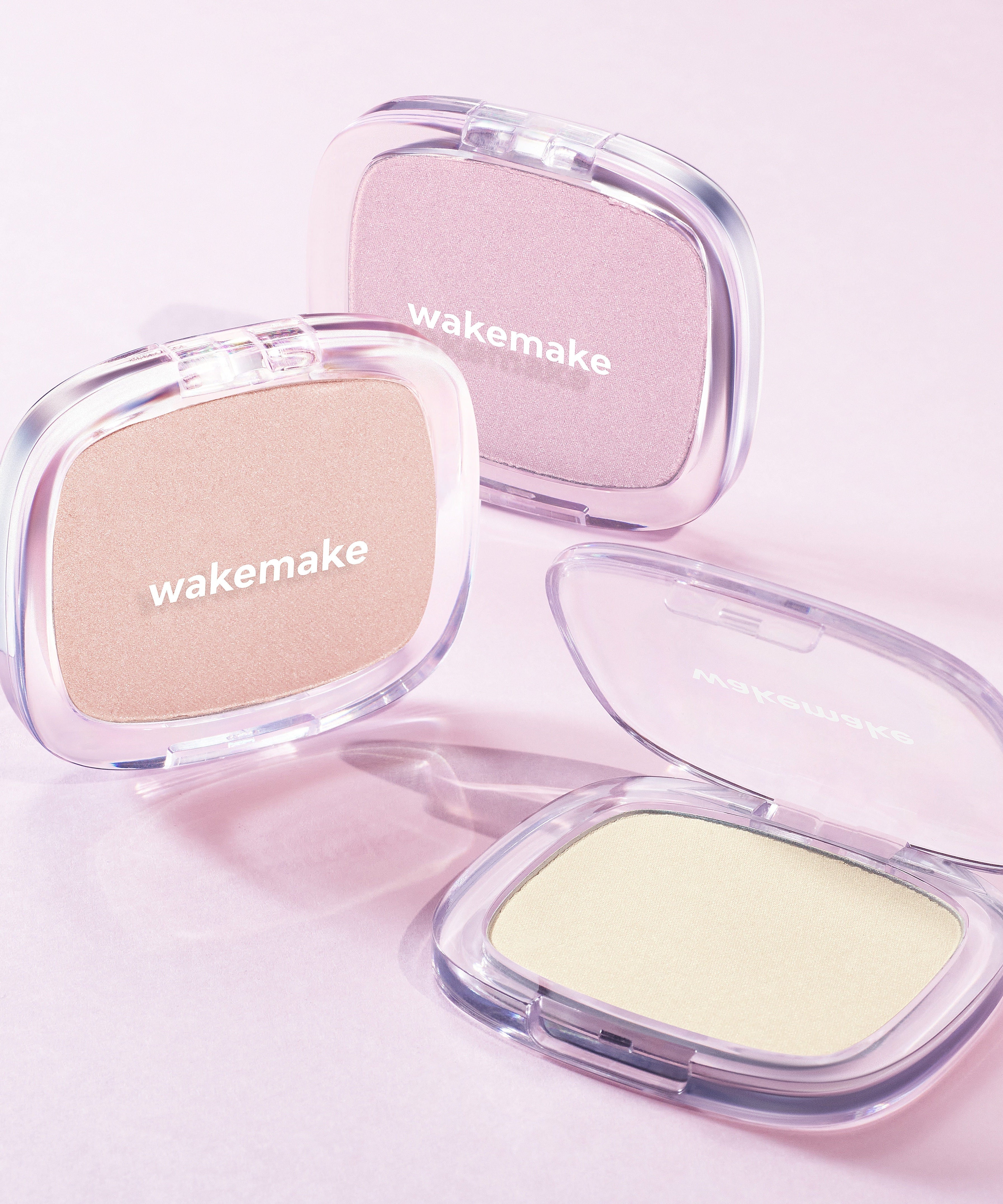 Three WAKE MAKE Sheer Breeze Highlighter 5g compacts in different shades on a pink background.