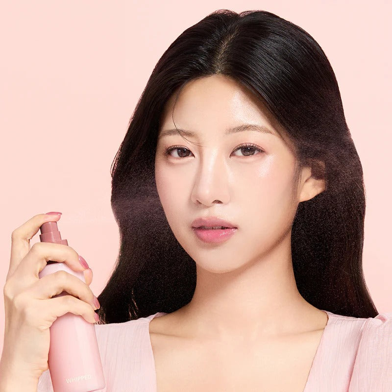 Model holding WHIPPED Butter Vegan Milk Foam Pack Mist 150ml product bottle against pink background.