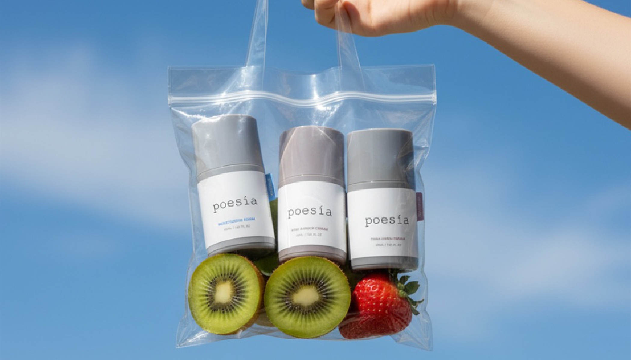 Hand holding clear bag with Poesia skincare products and fresh fruits against a blue sky background.